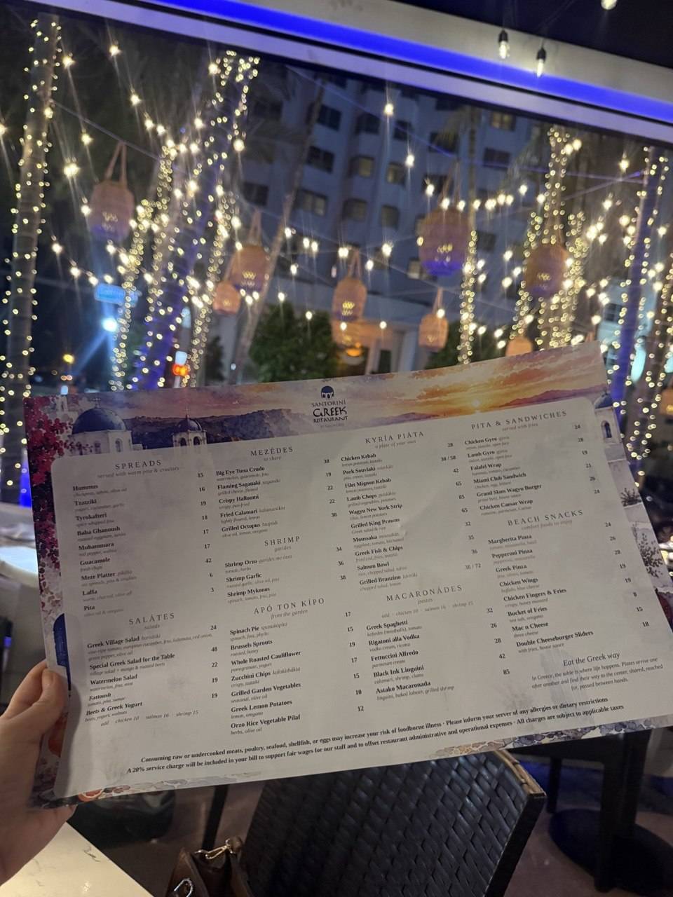 The Santorini by Georgios menu held beneath string lights and lanterns in the gardens of the Gale South Beach